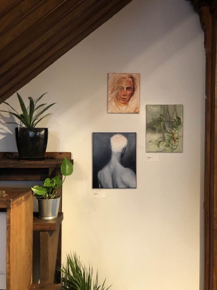 Three of Josh Iwata's paintings hung together on a gallery wall, beside potted plants on wood shelves